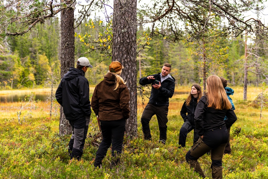 Lektion ute i skogen, med Jaktgymnasiet. En lärare står vid en gammeltall och berättar, elever lyssnar.